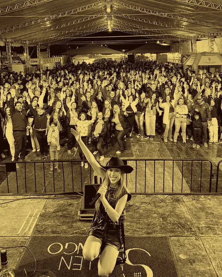 Rafaela Martins de chapéu no palco, com a plateia lotada atrás, em foto de tom sépia